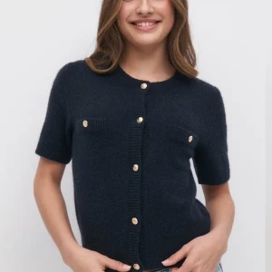 Svart kofta med korta ärmar och knappar - Säljer en stilren svart kofta med korta ärmar och dekorativa knappar framtill. Perfekt för en elegant och avslappnad look. Koftan har två fickor med knappdetaljer.