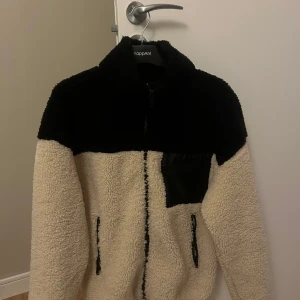 Beige/Svart  teddyjacka från Ginatricot - Mysig beige teddyjacka med svarta detaljer från Gina. Jackan har en dragkedja framtill och två fickor med dragkedja. Använd fåtalet gånger.