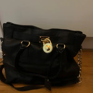 Svart skinnväska från Michael Kors - Säljer en elegant svart skinnväska från Michael Kors med gulddetaljer och kedjehandtag. Väskan har en dekorativ låsdetalj framtill och är perfekt för att ge en lyxig touch till din outfit. Den kommer med en skyddande dustbag.