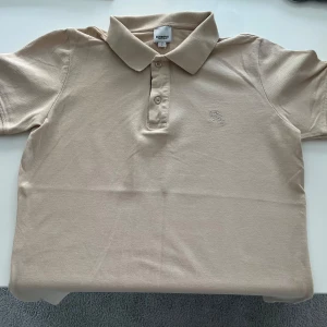 Beige pikétröja från Burberry - Säljer en stilren beige pikétröja från Burberry med klassisk krage och knappslå. Tröjan har korta ärmar och ett diskret broderat märke på bröstet. Insidan av knappslån har det ikoniska Burberry-mönstret. Perfekt för en avslappnad men elegant look. Använd 2-3 gånger.