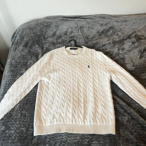 Vit stickad tröja från Ralph Lauren - Säljer en stilren vit stickad tröja från Ralph Lauren med kabelstickat mönster. Tröjan har långa ärmar och en liten broderad logotyp på bröstet. Perfekt för en klassisk och tidlös look.