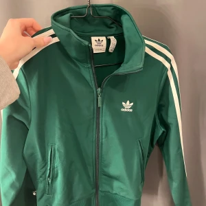 Grön träningsjacka från Adidas - Snygg grön träningsjacka från Adidas med klassiska vita ränder längs ärmarna. Jackan har dragkedja framtill och två fickor. Använd ett fåtal gånger 