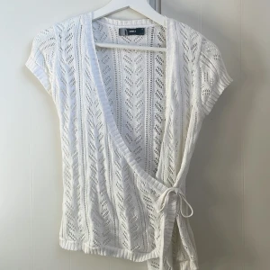 Vit omlottkofta från ONLY - Säljer en söt vit omlottkofta från ONLY med korta ärmar och knytband i midjan. Koftan har ett fint spetsmönster. Perfekt för lager-på-lager-stil!