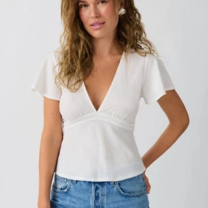 Vit v-ringad topp från Gina Tricot - Säljer en stilren vit topp från Gina Tricot med v-ringning och korta ärmar. Toppen har en knytning i ryggen. 💓💓