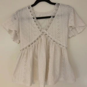 Vit blus med spetsdetaljer - Charmig vit blus med spetsdetaljer och korta ärmar. Blusen har ett omlottmönster framtill och är dekorerad med fina spetskanter. Perfekt för en somrig look🌸