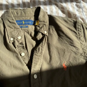 Olivgrön skjorta från Ralph Lauren - Säljer en olivgrön skjorta från Ralph Lauren med klassisk krage och knappar framtill. Skjortan har en broderad logotyp i rött på bröstet och långa ärmar. Perfekt för en stilren look! Hål vid handleden men lagat bra.
