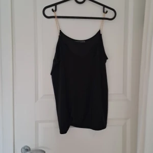 Svart linnetopp med kedjeband från Atmosphere - Snygg svart linnetopp från Atmosphere med unika kedjeband i guld. Perfekt för en stilren look. Toppen har en enkel design som gör den lätt att matcha med olika outfits.