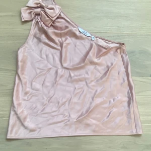 Beige oneshoulder topp med rosett - Jättefin rosa oneshoulder topp i satin med en stor rosett vid axeln. 💛Perfekt för en elegant look.❤️Strl 38 men passar S/M💕