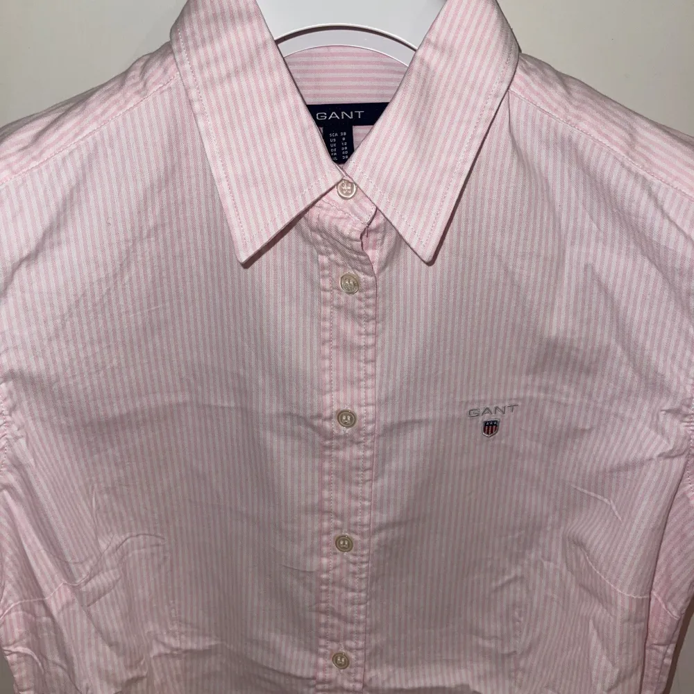 Säljer en stilren rosa randig skjorta från GANT med klassisk krage och knappar framtill. Skjortan har långa ärmar och ett diskret broderat märke på bröstet. Perfekt för en elegant look. Ordpris 1198kr mitt pris 279kr. Paidat.
