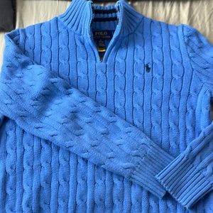 Blå stickad tröja från Ralph Lauren - Säljer en snygg blå stickad tröja från Ralph Lauren med kabelstickat mönster. Tröjan har en dragkedja vid halsen och långa ärmar. Perfekt för kyligare dagar!