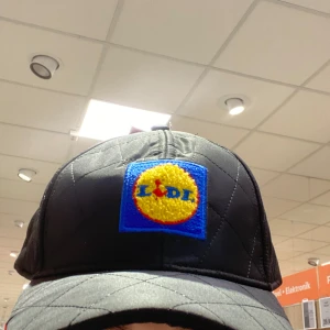 Svart keps med Lidl-logga - Snygg svart keps med broderad Lidl-logga i gult, rött och blått framtill. Perfekt för dig som vill ha en unik och rolig accessoar. Kepsen har en quiltad design som ger den en extra touch av stil.