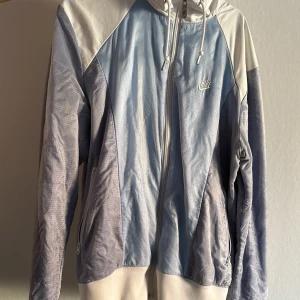 Vintage vindjacka från Nike - Säljer en stilren blå och vit vindjacka från Nike med dragkedja och huva. Jackan har långa ärmar och en stor Nike-logga på ryggen. Perfekt för en sportig look!