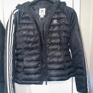 Svart dunjacka från Adidas - Säljer en snygg svart dunjacka från Adidas med klassiska vita ränder längs ärmarna. Jackan har en dragkedja framtill och en huva med dragsko 
