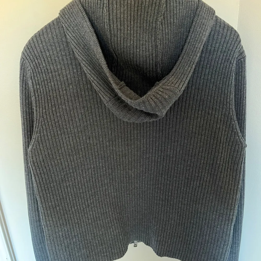 Snygg grå cardigan med dragkedja framtill. Tröjan har en ribbad design på ärmar och huva, vilket ger en stilren look. Perfekt för kyligare dagar. Skicket är super bra inga defekter alls. Den har aldrig använts. . Neuletakit & Villapaidat.
