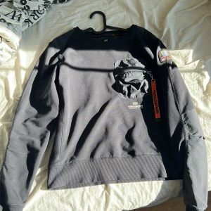 Svart fleece tröja från Parajumpers - Säljer en svart fleece tröja från Parajumpers med en praktisk ficka på bröstet och en snygg patch på ärmen. Tröjan har långa ärmar och en orange dragkedjedetalj. Perfekt för kyliga dagar!