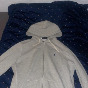 Grå hoodie från Ralph Lauren - Säljer en stilren grå hoodie från Ralph Lauren med dragkedja och klassisk logga på bröstet. Perfekt för en avslappnad look. Den har en justerbar huva med snören och långa ärmar.