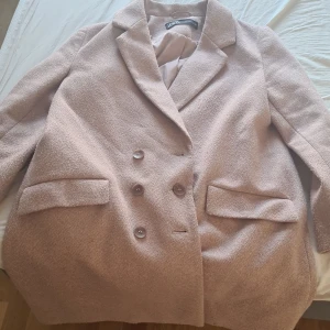 Rosa kappa från Zara - Säljer en stilren beige kappa från Zara med dubbelknäppning och klassisk krage. Kappan har två framfickor och en elegant passform som passar perfekt för höst och vår. Perfekt för att hålla stilen i kyligare väder!