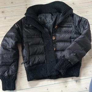 Svart dunjacka med stickade detaljer den har en defekt (se 2 bilden) - Snygg svart dunjacka med stickade muddar och krage. Jackan har knappar och dragkedja framtill samt ribbade detaljer som ger en cool look. Perfekt för kyligare dagar.