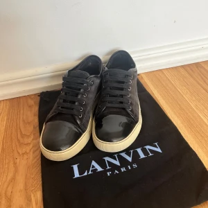 Svarta sneakers från Lanvin - Snygga svarta sneakers från Lanvin med en glansig tå och vita sulor. Skorna har snörning och en stilren design som passar perfekt för en trendig look. Perfekta för dig som vill ha en kombination av stil och komfort.dust bag medföljs 