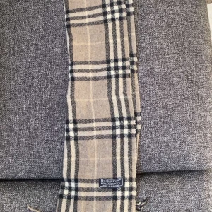 Beige rutig halsduk från Burberry - Säljer en klassisk beige rutig halsduk från Burberry. Den har ett tidlöst rutmönster i svart och vitt, Märkesetiketten är synlig på ena sidan. Perfekt accessoar för både vardag och speciella tillfällen.