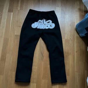 Svarta mjukisbyxor med tryck - Säljer ett par svarta mjukisbyxor med ett stort vitt tryck på framsidan. Byxorna har en elastisk midja och en avslappnad passform, perfekta för en bekväm stil. Trycket har en cool design med text och stjärnor. 