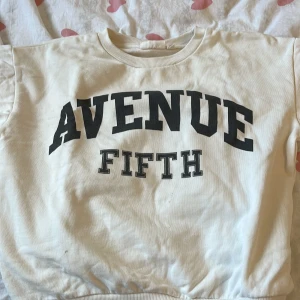 Vit sweatshirt från KappAhl - Säljer en vit sweatshirt från KappAhl med trycket 'Avenue Fifth' i svart på framsidan. Tröjan har en rund halsringning och ribbade muddar vid ärmslut och nederkant. 