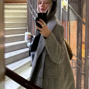 Grå ullkappa - Snygg grå kappa i ull med klassisk design och breda slag. Perfekt för kyliga dagar med sin långa passform och stilrena look. Kappan har knappar framtill och är helfodrad för extra komfort.