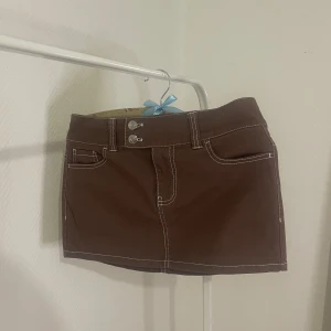 Brun kortkjol med vita sömmar - Snygg brun kortkjol med kontrasterande vita sömmar och klassisk jeansstil. Kjolen har fickor och bälteshällor. Låg midjan som är väldigt trendig just nu.