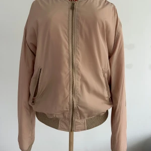 Beige bomberjacka NY - Snygg beige bomberjacka från DM Retro med en cool orange quiltad insida. Jackan har dragkedja framtill och på ena ärmen samt ribbade muddar vid ärmslut och nederkant. Perfekt för en stilren look! Aldrig använd. Stl L men liten i storleken.