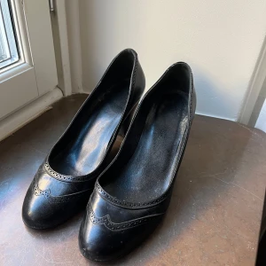 Svarta pumps med klack - Eleganta svarta pumps i skinn med dekorativa detaljer och en bekväm klack. Perfekta för en stilren look.
