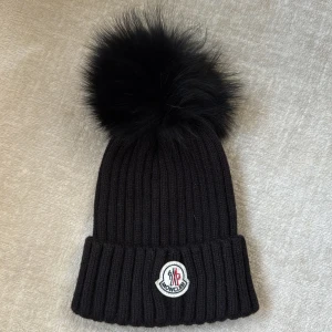 Svart ribbad mössa med pälsboll från Moncler - Snygg svart ribbad mössa från Moncler med en stor pälsboll på toppen. Mössan har en broderad logotyp framtill och är perfekt för kyliga dagar. Knappt använd då jag fick den i present.