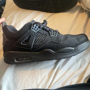 Svarta sneakers från Nike - Säljer ett par svarta jordan 4 black cat sneakers med ikonisk design. Skorna har snörning och en synlig Air-enhet i sulan för extra komfort. Ovandelen är i mocka med nätpaneler för ventilation. Perfekta för både stil och funktion.