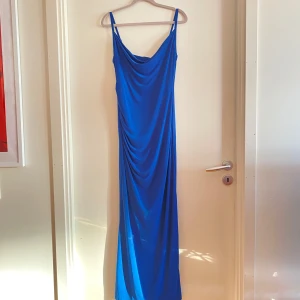 Blå långklänning med smala axelband - Elegant blå långklänning med smala axelband och draperad design. Klänningen har en figurnära passform och är perfekt för speciella tillfällen. Den är tillverkad i ett mjukt och följsamt material som ger en smickrande silhuett.