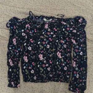 Blommig offshoulder blus från H&M - Säljer en söt blommig offshoulder blus från H&M. Blusen har ett färgglatt mönster med rosa, blå och vita blommor på en mörk bakgrund. Den har långa ärmar och en elastisk kant vid axlarna för en bekväm passform. Skriv gärna om du vill ha fler bilder eller exakt mått på något speciellt 