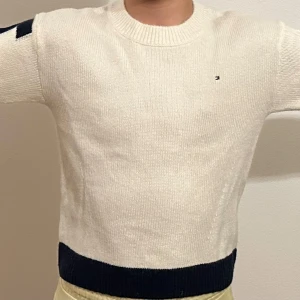 Vit stickad tröja från Tommy Hilfiger - Säljer en stilren vit stickad tröja från Tommy Hilfiger med mörkblå och röda detaljer. Tröjan har långa ärmar och en ribbad kant vid ärmslut och nederkant. Perfekt för en klassisk look.