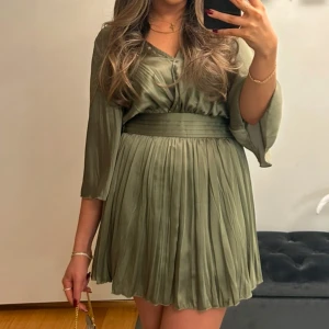 Grön klänning med plisserad kjol - Snygg grön klänning med plisserad kjol och trekvartsärmar. Plagget har ”shorts” i sig som en jumpsuit men det ser ut som en klänning Den har en markerad midja och ett vackert fall. Perfekt för en stilren look.