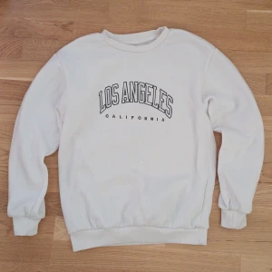 Vit sweatshirt med Los Angeles-tryck - Säljer en vit sweatshirt med texten 'Los Angeles California' tryckt på framsidan. Tröjan har långa ärmar och ribbade muddar vid ärmslut och nederkant. Perfekt för en avslappnad stil.