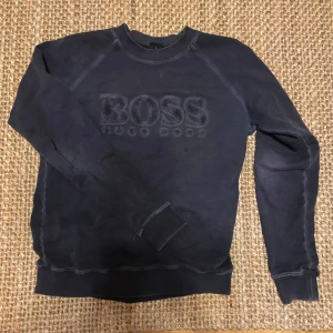 Marinblå Hugo Boss tröja - Säljer denna mörkblåa sweathsirten från Hugo Boss. Den är ganska sliten och urtvättad och använder inte den så vill bli av med den, sitter som M/L. Kom privat för fler frågor!