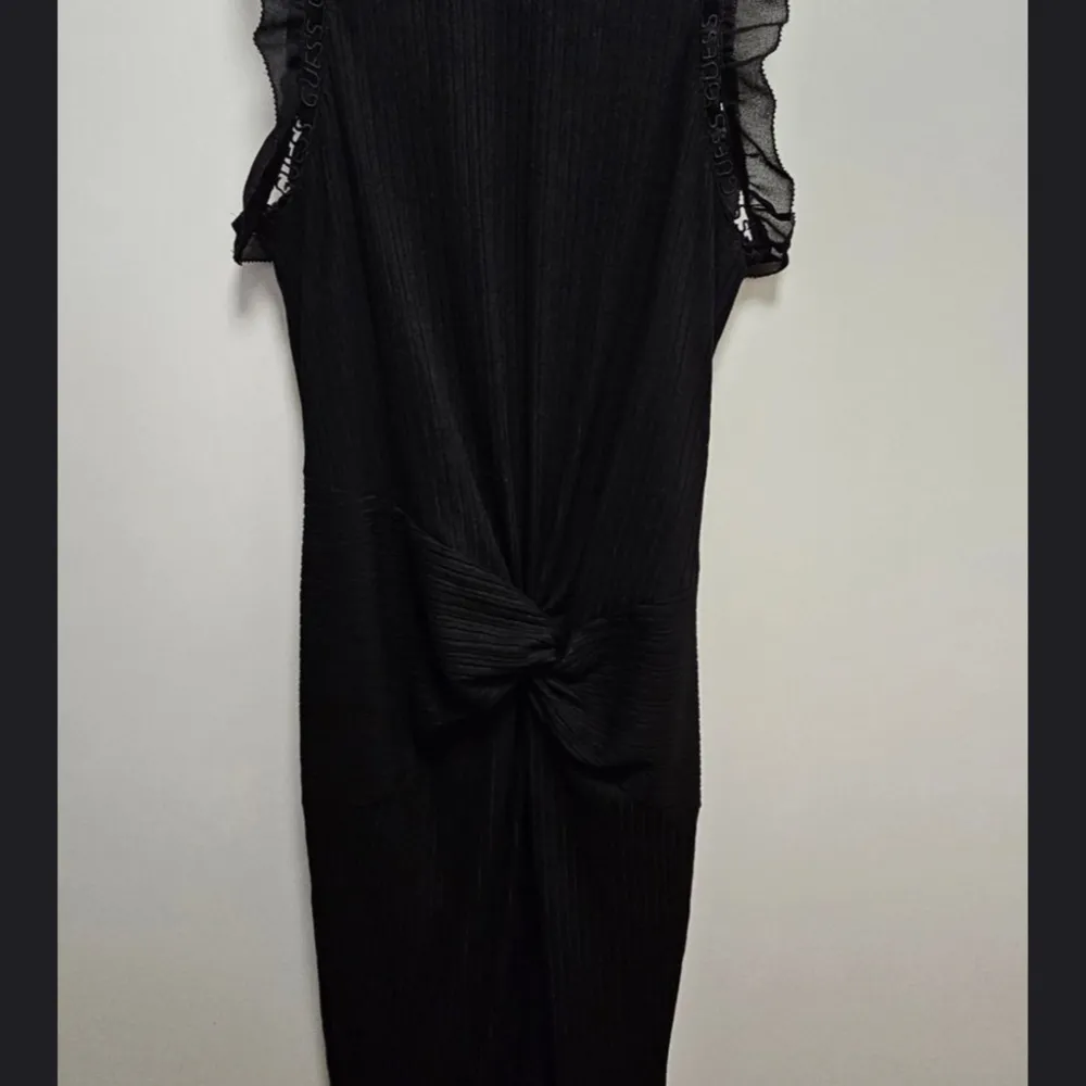 Elegant svart klänning från Guess med ribbad textur och volangdetaljer vid ärmarna. Klänningen har en knytning framtill och en diskret dragkedja bak. Perfekt för en stilren look.. Mekot.