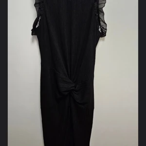 Svart klänning från Guess - Elegant svart klänning från Guess med ribbad textur och volangdetaljer vid ärmarna. Klänningen har en knytning framtill och en diskret dragkedja bak. Perfekt för en stilren look.