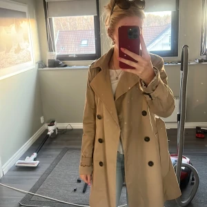 Beige trenchcoat - Snygg beige trenchcoat med klassisk design och knappar framtill. Jackan har ett bälte i midjan och långa ärmar. Perfekt för vår och höst.