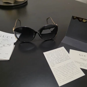 Svarta solglasögon från Gucci - Snygga svarta solglasögon från Gucci med ovala linser och guldfärgade detaljer. Skalmarna har det ikoniska Gucci-emblemet och ränder i grönt och rött. Kommer med verifikationspapper och kvitto