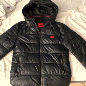 Svart pufferjacka från Hugo Boss - Säljer en stilren svart pufferjacka från Hugo Boss med huva och dragkedja. Jackan har en quiltad design och är perfekt för kyligare väder. Den har praktiska fickor med dragkedjor och en snygg röd detalj på insidan. Jackan är köpt 2023 för ordinarie pris på 3600kr, priset på jackan ligger i nuläget på 2600kr. Kvitto och paketering ingår inte.