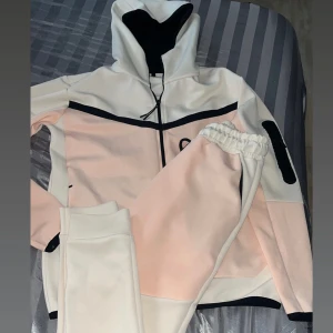 Vit och rosa kofta med byxor - Säljer en snygg vit och rosa kofta med matchande byxor. koftan har dragkedja och svarta detaljer, inklusive en huva med snörning. Byxorna har elastisk midja och svarta snören. Perfekt för en avslappnad stil.