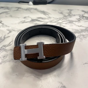 Brunt skärp med silverfärgat H-spänne - Snyggt brunt skärp med ett iögonfallande silverfärgat H-spänne. Perfekt för att ge en stilren touch till din outfit. Skärpet är tillverkat i skinn och har en klassisk design som passar till många olika stilar. Obs! Är en tjejmodell så något mindre än herr modell!