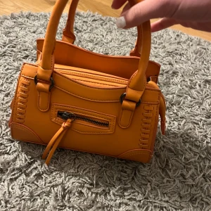 Orange handväska med dragkedja - Säljer en snygg orange handväska med dekorativa sömmar och en praktisk dragkedja framtill. Väskan har två handtag och en kompakt design som gör den perfekt för att bära med sig det viktigaste. Perfekt för att liva upp din outfit med en färgklick!