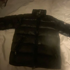 Svart dunjacka från Moncler - Säljer en snygg svart dunjacka från Moncler. Jackan har en hög krage och dragkedja framtill. Den är quiltad och har långa ärmar med tryckknappar. Perfekt för kyliga dagar! Dock har den ingen luva så säljer den billigare. Den är köpt för 1600kr