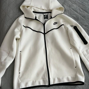 Vit hoodie från Nike - Säljer en stilren vit hoodie från Nike med svarta detaljer och dragkedja. Jackan har en huva och långa ärmar, perfekt för en sportig look. Den har även en praktisk ficka med dragkedja på ärmen.