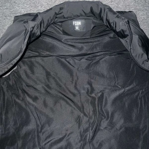 Svart dunväst från FSBN - Snygg svart dunväst från FSBN med dragkedja framtill. Västen har ett mönster med texten 'CHAOTIC' och är perfekt för kyligare dagar. Den har en modern look och passar bra till olika outfits.