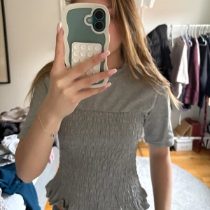 Grå smockad topp med kort ärm - Säljer en ljusgrå topp med smockad midja och korta ärmar. Toppen har en figurnära passform upptill och volangliknande avslut nedtill. Perfekt för en avslappnad och trendig look.